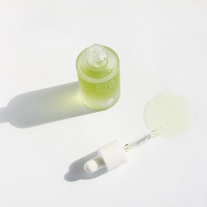 Whamisa ﻿Organic Flowers Facial Oil Refresh bottled in a semi-transparent bottle with lime green content inside. The cap is off and it works as a dropper. The content is spilled on the ground to highlight the lime green color of the content.